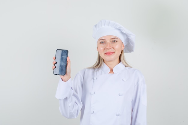 Vrouwelijke chef-kok die smartphone toont en in wit uniform glimlacht en vrolijk kijkt.