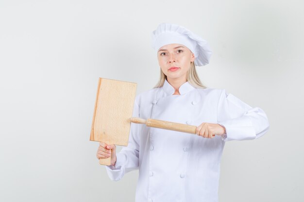 Vrouwelijke chef-kok die snijplank en deegroller in wit uniform houdt en serieus kijkt