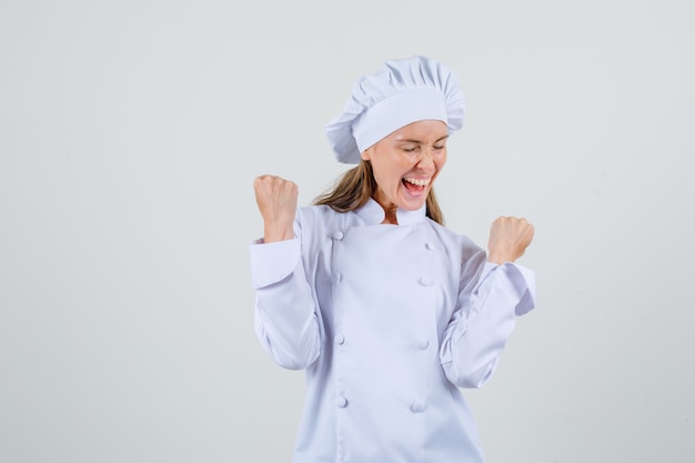 Vrouwelijke chef-kok die winnaargebaar in wit uniform toont en gelukkig kijkt. vooraanzicht.