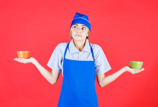 Vrouwelijke chef-kok in blauwe schort die oranje en groene keramische noedelbekers vasthoudt en nadenkt over hoe ze te gebruiken