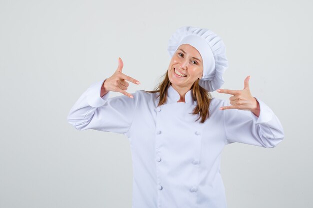 Vrouwelijke chef-kok in wit uniform met 'ik hou van jou'-gebaar en ziet er vrolijk uit