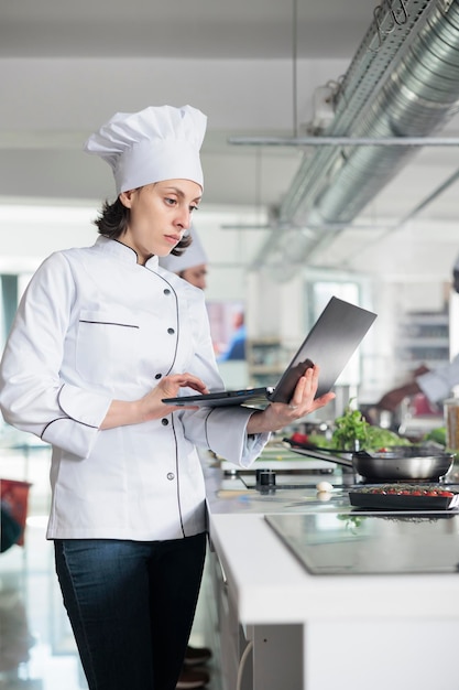 Vrouwelijke chef-kok met laptop brainstormen garnituur ideeën voor gastronomisch gerecht recept terwijl je in de keuken van het restaurant staat. Professionele werknemer in de voedingsindustrie met computer op zoek naar gastronomische maaltijd.