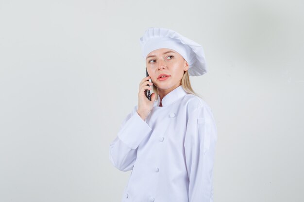 Vrouwelijke chef-kok praten over smartphone en opzij kijken in wit uniform.