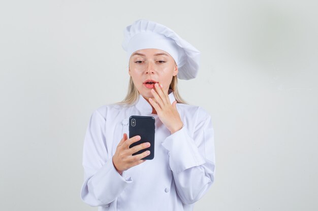 Vrouwelijke chef-kok smartphone in wit uniform houden en verbaasd kijken