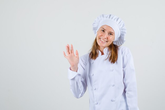 Vrouwelijke chef-kok zwaaiende hand om hallo in wit uniform te zeggen en er blij uit te zien. vooraanzicht.