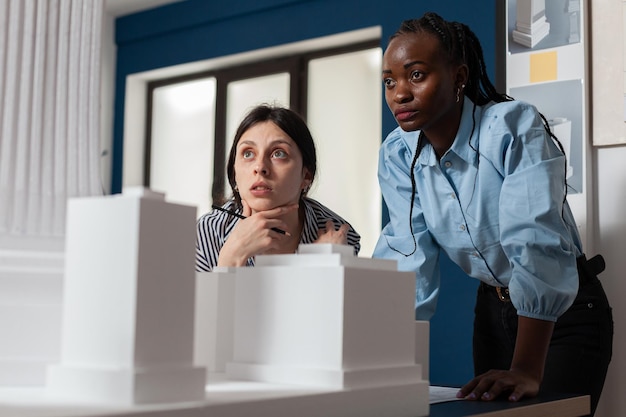 Vrouwelijke collega's van projectingenieurs kijken geïnspireerd voor 3D-woonmaquette in architectonisch modern kantoor. Geconcentreerd professioneel architectenteam dat wolkenkrabber wit schuimmodel inspecteert.