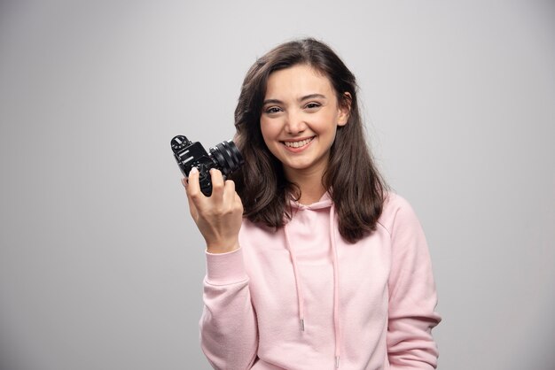 Vrouwelijke fotograaf die met camera op grijze muur glimlacht.