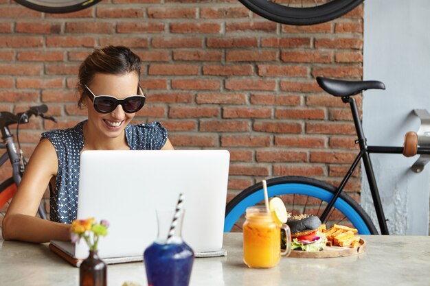 Vrouwelijke freelancer met gelukkige glimlach die op afstand aan laptop werkt. Succesvolle foodblogger die een nieuw bericht op haar blog typt