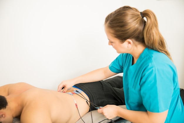 Vrouwelijke fysiotherapeut die elektroden op de klant plaatst voor behandeling van de lage rugspieren in de kliniek