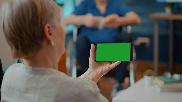 Vrouwelijke gepensioneerde m/v kijken naar horizontaal groen scherm op mobiele telefoon. oude vrouw met krukken met smartphone met lege chromakey en geïsoleerde kopie ruimte achtergrond. mock-up sjabloon