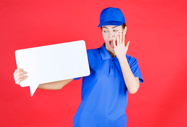 Vrouwelijke gids in blauw uniform met een wit rechthoekig infobord en ziet er doodsbang en verrast uit.