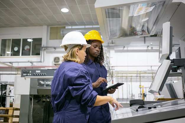 Vrouwelijke industriële werknemer lesgeven collega om machine te bedienen, wijzend op de besturingskaart, met behulp van Tablet PC