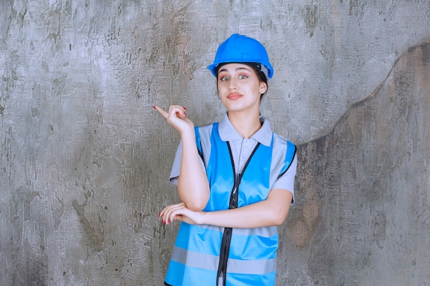 Vrouwelijke ingenieur die een blauwe helm en uitrusting draagt en met emoties naar iets aan de linkerkant wijst