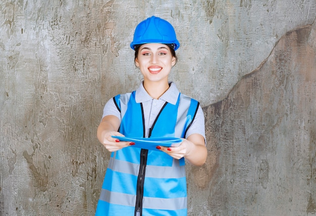 Vrouwelijke ingenieur in blauw uniform en helm die een blauwe rapportmap vasthoudt en ter verificatie geeft