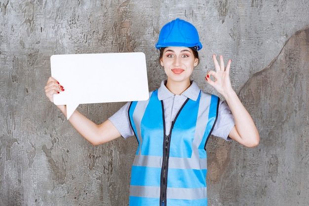 Gratis foto vrouwelijke ingenieur in blauw uniform en helm die een leeg rechthoekig infobord houdt en plezierteken toont.