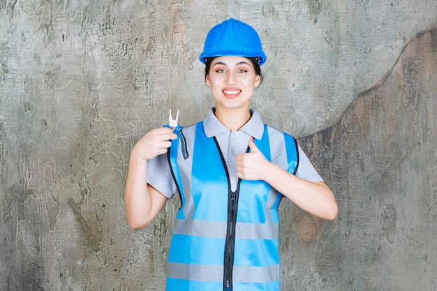 Vrouwelijke ingenieur in blauw uniform en helm die een metalen tang vasthoudt voor reparatie en een positief handteken toont