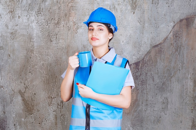 Vrouwelijke ingenieur in blauw uniform en helm met een blauwe theekop en een blauwe rapportmap.