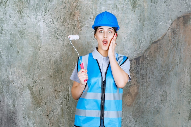 Vrouwelijke ingenieur in blauw uniform en helm met een trimrol om te schilderen en ziet er bang en doodsbang uit. Premium Foto