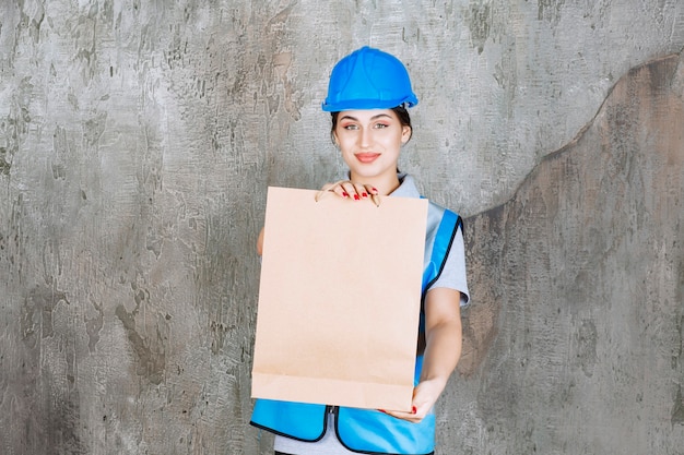 Vrouwelijke ingenieur in blauwe helm en uitrusting met een kartonnen boodschappentas.