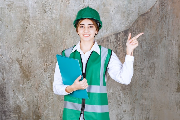 Vrouwelijke ingenieur in groen uniform en helm die een groene projectmap vasthoudt en naar de rechterkant wijst.