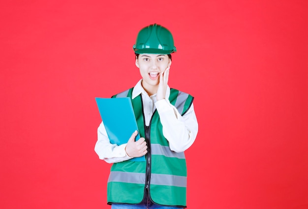 Vrouwelijke ingenieur in groen uniform met een blauwe map en ziet er bang en doodsbang uit