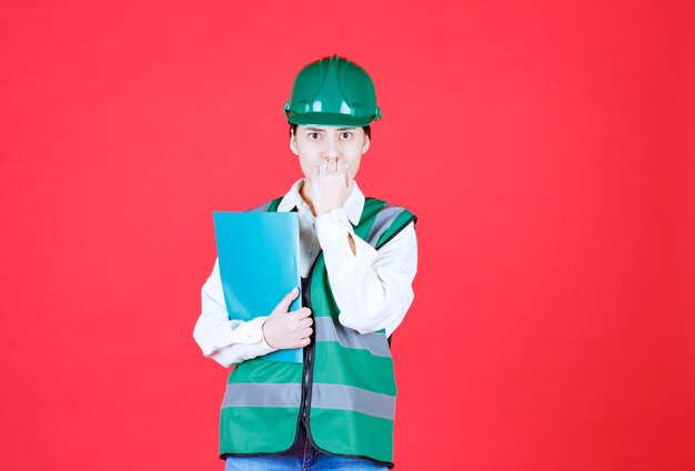 Vrouwelijke ingenieur in groen uniform met een blauwe map en ziet er bang en doodsbang uit