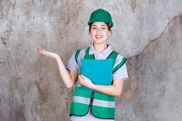 Vrouwelijke ingenieur in groene helm die een blauwe map vasthoudt en naar iemand in de buurt wijst.