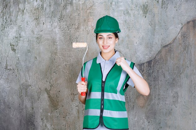 Vrouwelijke ingenieur in groene helm die een trimrol vasthoudt voor muurschildering en een positief handteken toont.