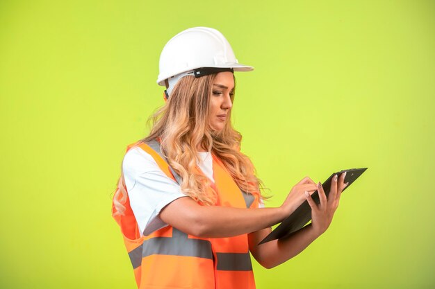 Vrouwelijke ingenieur in witte helm en versnelling die de controlelijst vasthoudt en aantekeningen maakt.
