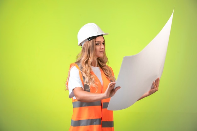 Vrouwelijke ingenieur in witte helm en vistuig die het bouwplan controleert en het leest