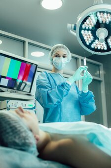 Vrouwelijke jonge medische werker met een spuit in handen die onder de chirurgische lamp staat en naar de patiënt kijkt