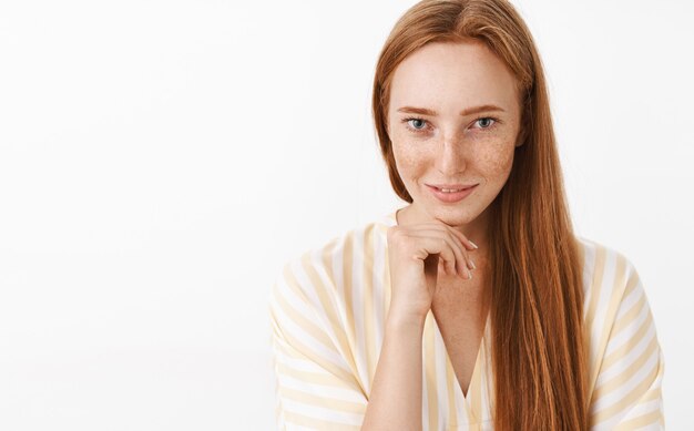 Vrouwelijke knappe flirterige roodharige vrouw met sproeten in schattige v-hals jurk met hand op de kaak en lachend met sensuele verleidelijke blik