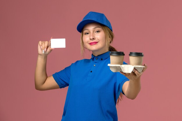 Vrouwelijke koerier in blauw uniform bedrijf levering kopjes koffie en witte kaart met glimlach op roze, service uniforme levering