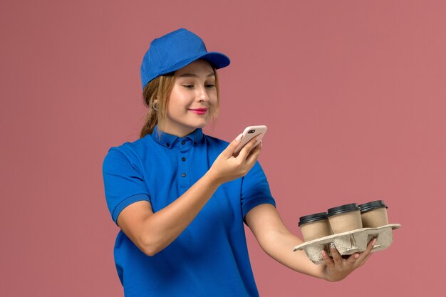 vrouwelijke koerier in blauw uniform die een foto neemt van bruine kopjes koffie op lichtroze, werkdienstuniforme levering