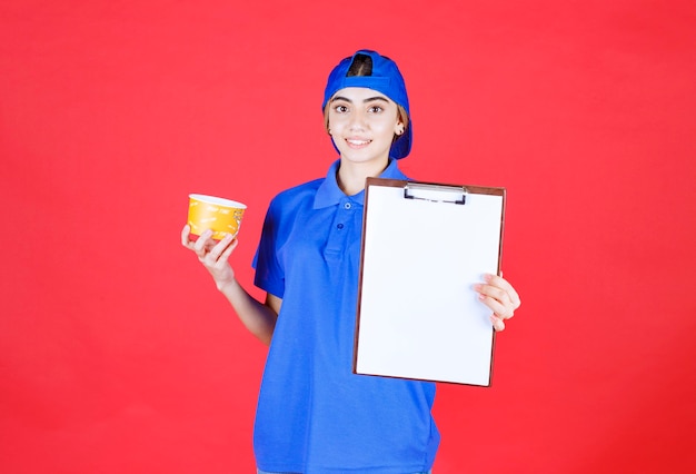 Vrouwelijke koerier in blauw uniform die een gele noedelsbeker vasthoudt en de takenlijst ter ondertekening presenteert.