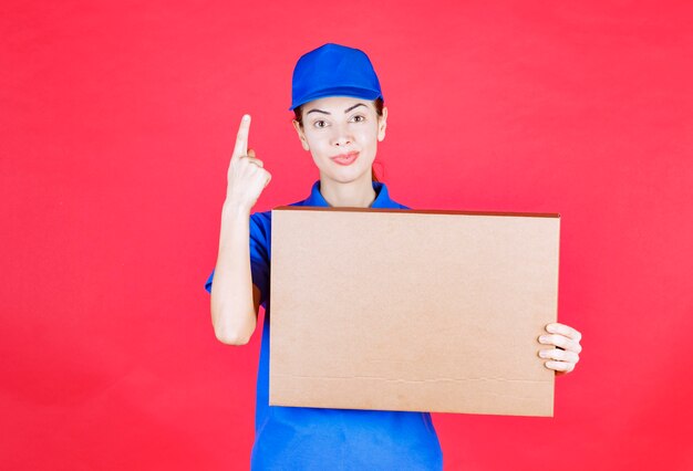 Vrouwelijke koerier in blauw uniform die een kartonnen afhaalpizzadoos vasthoudt en verward denkt of kijkt.