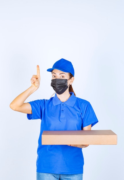 Vrouwelijke koerier in blauw uniform en gezichtsmasker met een kartonnen doos en ergens naar wijzend.