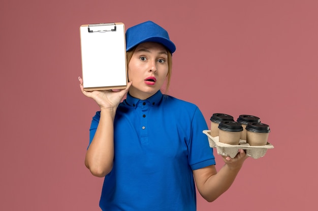 Gratis foto vrouwelijke koerier in blauw uniform met blocnote samen met bruine bezorgkopjes koffie op lichtroze, uniforme bezorging van de werkdienst