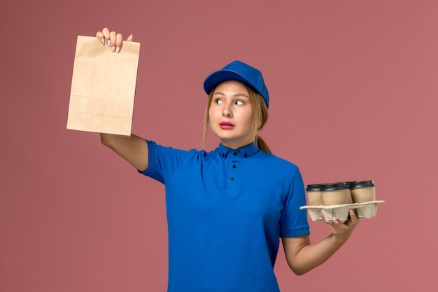 Vrouwelijke koerier in blauw uniform met voedselpakket en bruine levering kopjes koffie op roze, service uniforme levering baan