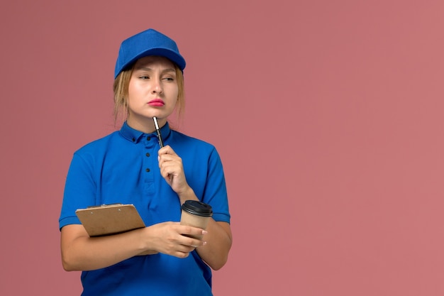 vrouwelijke koerier in blauw uniform poseren bedrijf kopje koffie en blocnote denken op roze, service uniform levering meisje