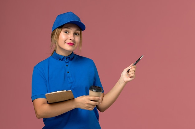 vrouwelijke koerier in blauw uniform poseren bedrijf kopje koffie en blocnote op roze, service uniforme levering meisje werknemer kleurenfoto
