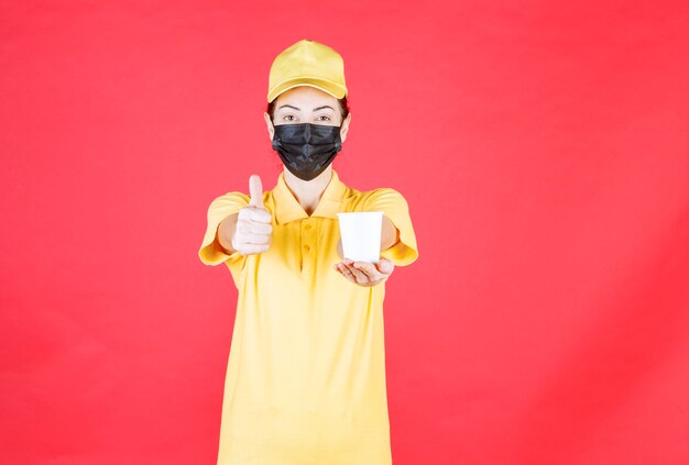 Vrouwelijke koerier in geel uniform en zwart masker met een afhaalbeker en genietend van de smaak