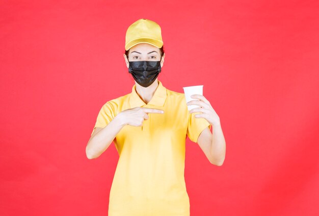 Vrouwelijke koerier in geel uniform en zwart masker met een afhaalbeker