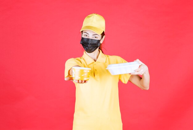 Vrouwelijke koerier in geel uniform en zwart masker met een afhaalpakket en een noedelbeker