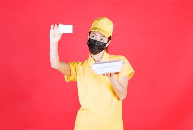 Vrouwelijke koerier in geel uniform en zwart masker met een afhaalpakket en presenteert haar visitekaartje