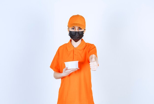 Vrouwelijke koerier in geel uniform en zwart masker met een plastic beker en positief handteken tonen.