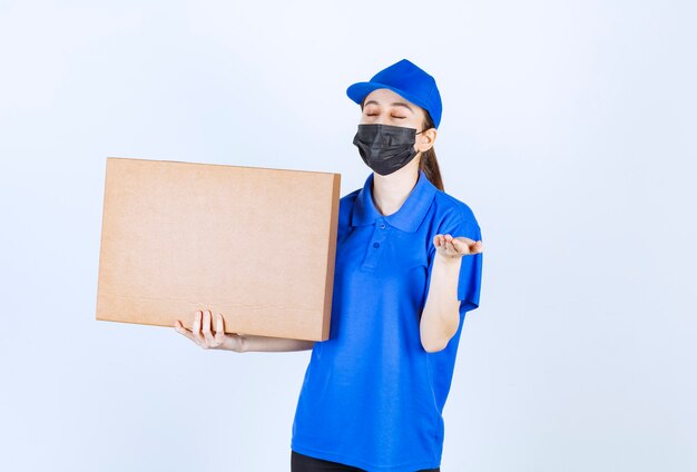 Vrouwelijke koerier in masker en blauw uniform die een groot kartonnen pakket vasthoudt en het product ruikt