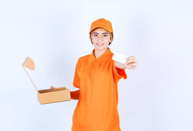 Vrouwelijke koerier in oranje uniform houdt een open kartonnen doos vast en stelt haar visitekaartje in op de klant