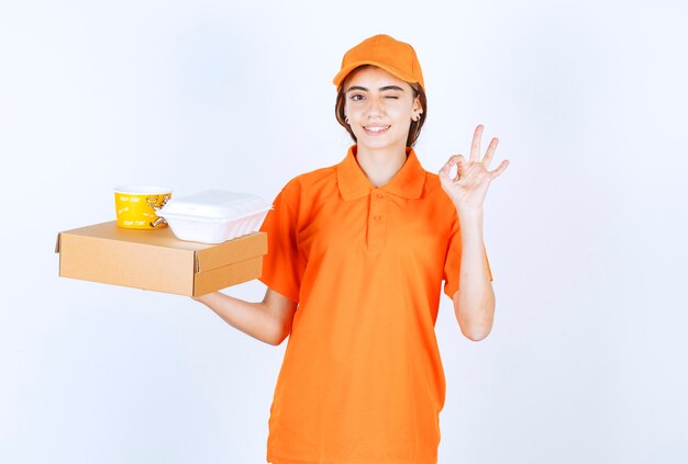 Vrouwelijke koerier in oranje uniform met gele en witte afhaaldozen met een kartonnen pakket en genietend van de smaak