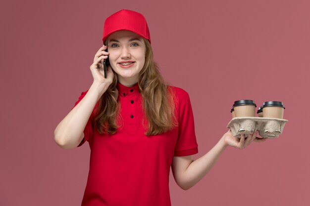 vrouwelijke koerier in rood uniform bedrijf bezorgkoffie praten aan de telefoon met glimlach op roze, baan uniforme werknemer dienstverlening